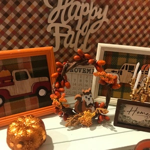 May include: A fall-themed vignette with a wooden sign that reads "Happy Fall", a framed picture of a truck with pumpkins, a framed calendar with the month of November, a wreath with a squirrel, a candlestick with white candles, and a framed sign that reads "Home".