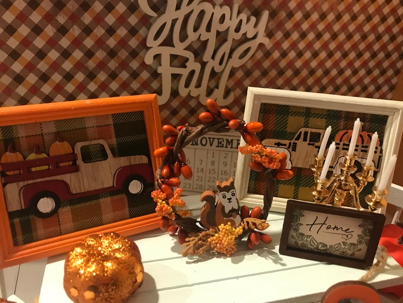 May include: A fall-themed vignette with a wooden sign that reads "Happy Fall", a framed picture of a truck with pumpkins, a framed calendar with the month of November, a wreath with a squirrel, a candlestick with white candles, and a framed sign that reads "Home".