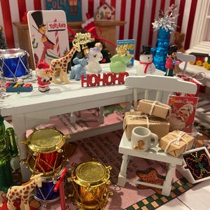 May include: A miniature Christmas scene with a white table adorned with festive figurines, including a giraffe, snowman, and Santa Claus. Miniature drums, wrapped gifts, and a toy train set add to the holiday theme. The word "HOHOHO" is displayed.