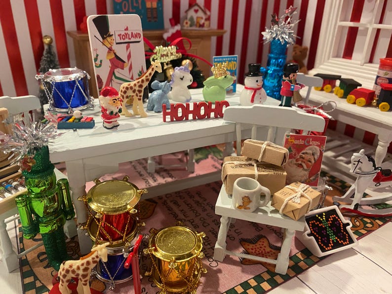 May include: A miniature Christmas scene with a white table adorned with festive figurines, including a giraffe, snowman, and Santa Claus. Miniature drums, wrapped gifts, and a toy train set add to the holiday theme. The word "HOHOHO" is displayed.