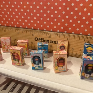 Op de afbeelding: Een verzameling miniatuur speelgoeddozen, elk met een andere karakterillustratie. De dozen zijn voornamelijk roze, blauw en geel, met gedetailleerde illustraties. Een houten liniaal staat op de achtergrond en de dozen staan op een wit oppervlak.