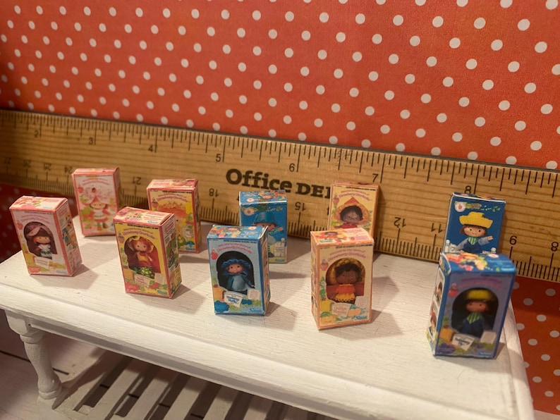 Op de afbeelding: Een verzameling miniatuur speelgoeddozen, elk met een andere karakterillustratie. De dozen zijn voornamelijk roze, blauw en geel, met gedetailleerde illustraties. Een houten liniaal staat op de achtergrond en de dozen staan op een wit oppervlak.