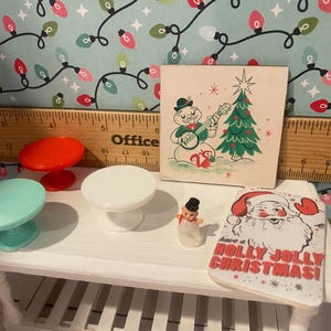 May include: A white table with three miniature tables, a snowman figurine, and a Christmas card that says "Have a Holly Jolly Christmas!" The miniature tables are red, white, and light blue. The card is on a white surface with a red and white Santa Claus illustration.