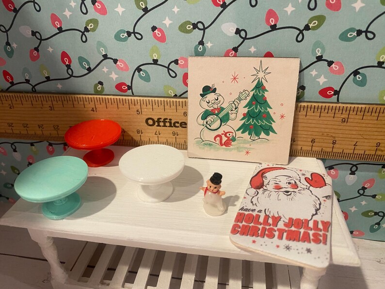 May include: A white table with three miniature tables, a snowman figurine, and a Christmas card that says "Have a Holly Jolly Christmas!" The miniature tables are red, white, and light blue. The card is on a white surface with a red and white Santa Claus illustration.