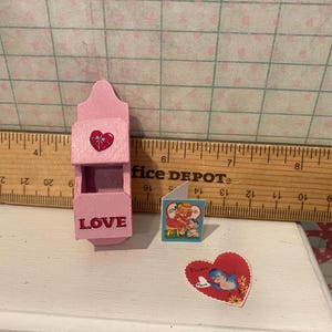 Może przedstawiać: Miniaturowa r&oacute;żowa drewniana skrzynka pocztowa z czerwonym sercem i napisem "LOVE" z przodu. Obecna jest r&oacute;wnież mała kartka z życzeniami i naklejka w kształcie serca z niebieskim ptakiem. Tematem jest Walentynki.