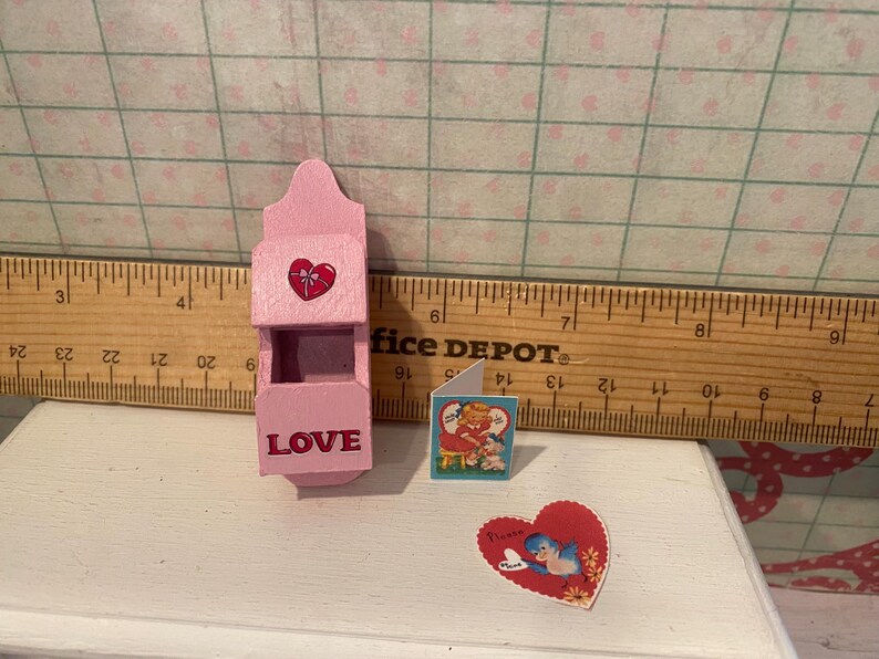 Może przedstawiać: Miniaturowa r&oacute;żowa drewniana skrzynka pocztowa z czerwonym sercem i napisem "LOVE" z przodu. Obecna jest r&oacute;wnież mała kartka z życzeniami i naklejka w kształcie serca z niebieskim ptakiem. Tematem jest Walentynki.