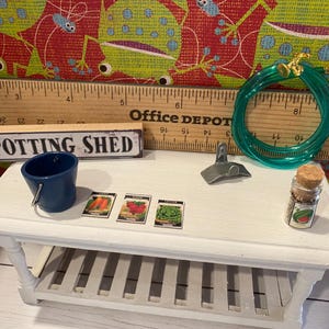 May include: A miniature potting shed scene featuring a blue bucket, seed packets, a green hose, and a small jar. The scene is set on a white table with a wooden ruler and a sign that reads "POTTING SHED".