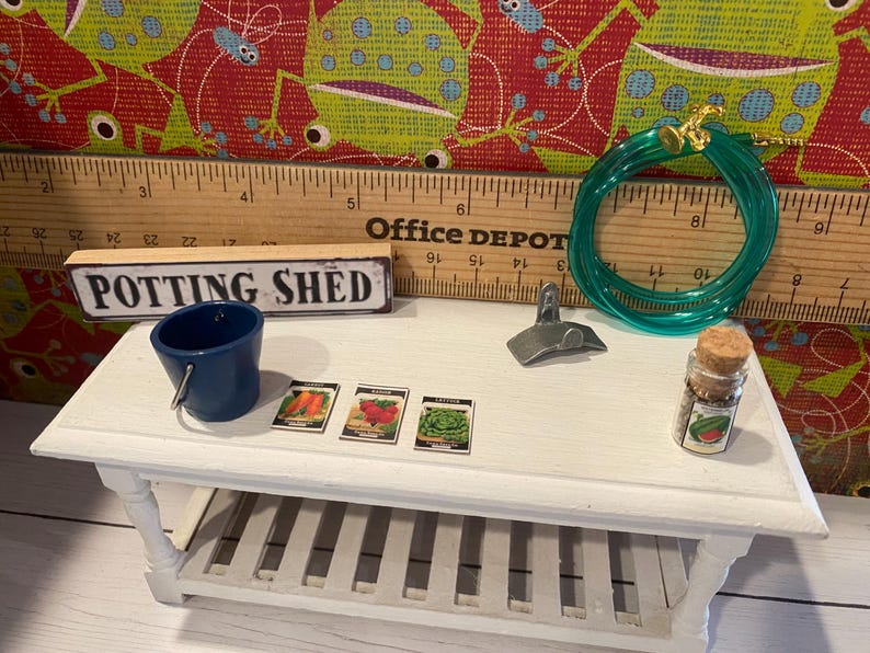 May include: A miniature potting shed scene featuring a blue bucket, seed packets, a green hose, and a small jar. The scene is set on a white table with a wooden ruler and a sign that reads "POTTING SHED".