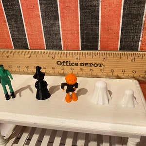 May include: Five Halloween-themed figurines: a green Frankenstein, a black witch, an orange pumpkin, and two white ghosts. The figurines are arranged on a white table with a wooden ruler behind them. The ruler is labeled "Office DEPOT".