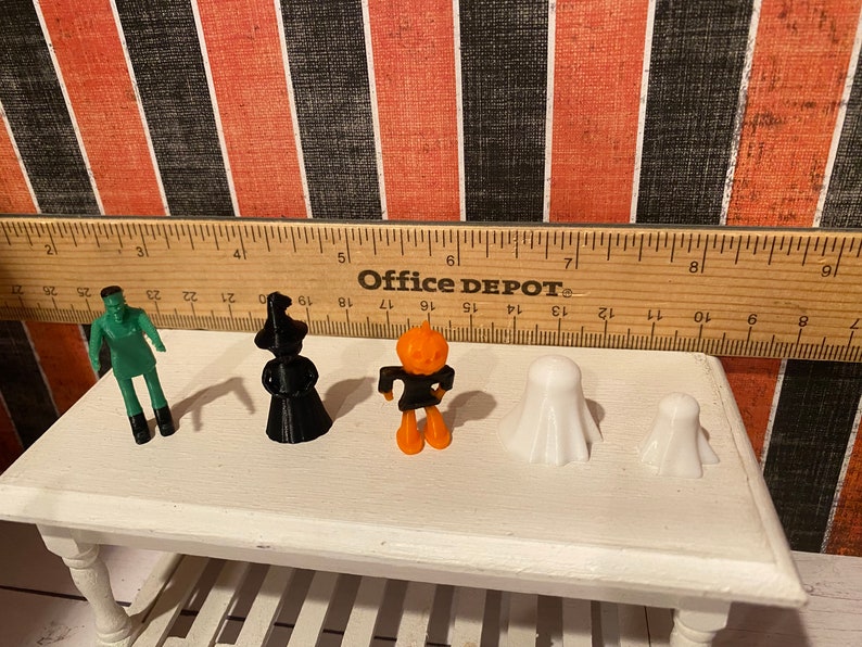 May include: Five Halloween-themed figurines: a green Frankenstein, a black witch, an orange pumpkin, and two white ghosts. The figurines are arranged on a white table with a wooden ruler behind them. The ruler is labeled "Office DEPOT".
