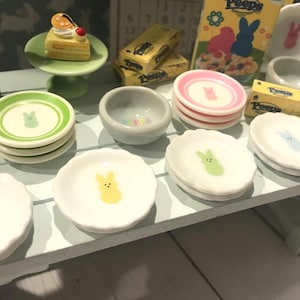 Puede incluir: Platos y cuencos de cerámica en miniatura con temática de Pascua con diseños de conejitos en varios colores. Incluye platos pequeños con conejitos verdes, rosas, amarillos y azules, y un pastel pequeño con una cereza.
