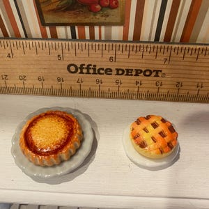 May include: Miniature baked goods, including a round pie with a lattice crust and a tart with a fluted edge, sit on small plates. The Office Depot ruler in the background provides scale. The pie and tart are golden brown and orange.