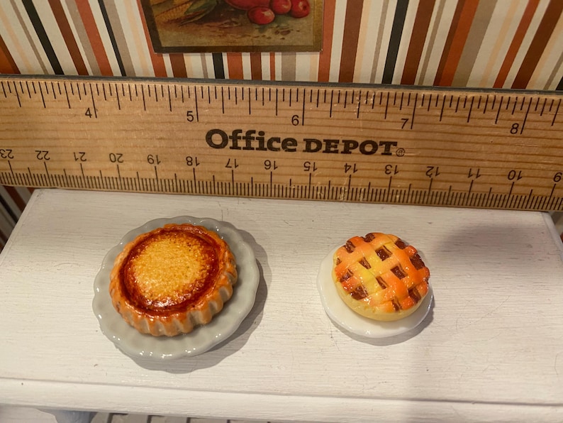 May include: Miniature baked goods, including a round pie with a lattice crust and a tart with a fluted edge, sit on small plates. The Office Depot ruler in the background provides scale. The pie and tart are golden brown and orange.