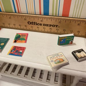 May include: A collection of miniature books on a white table, including titles like "Goodnight Moon" and "Guess How Much I Love You." A wooden ruler is in the background. The books are colorful and feature illustrations.