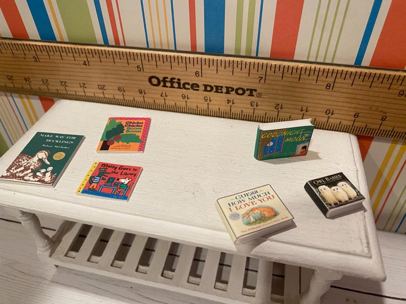 May include: A collection of miniature books on a white table, including titles like "Goodnight Moon" and "Guess How Much I Love You." A wooden ruler is in the background. The books are colorful and feature illustrations.