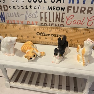 May include: Four ceramic cat figurines on a white table. The cats are in various poses, including sitting, lying down, and stretching. The cats are white, orange, black, and white, and orange and white.