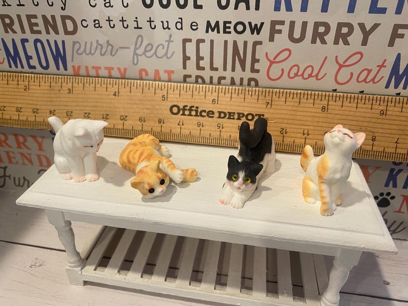 May include: Four ceramic cat figurines on a white table. The cats are in various poses, including sitting, lying down, and stretching. The cats are white, orange, black, and white, and orange and white.