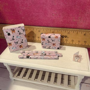 K&ouml;nnte beinhalten: Ein Miniatur-Set bestehend aus einer rosa Papiert&uuml;te, einer kleinen rechteckigen Schachtel, drei d&uuml;nnen zylindrischen R&ouml;hren und einer winzigen quadratischen Gru&szlig;karte, alle mit einem wiederkehrenden Katzenmuster verziert. Ein Holzlineal ist im Hintergrund sichtbar.