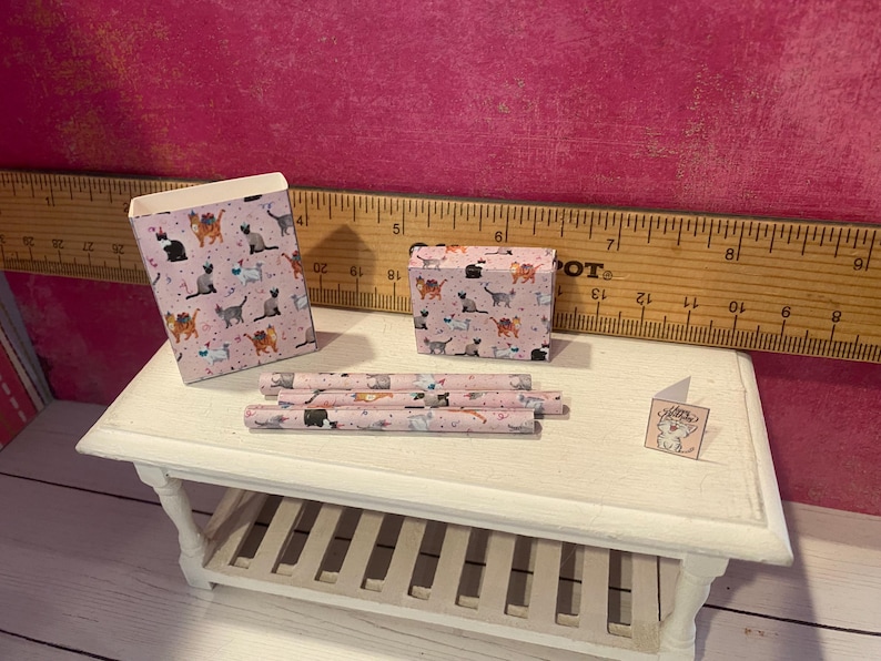 K&ouml;nnte beinhalten: Ein Miniatur-Set bestehend aus einer rosa Papiert&uuml;te, einer kleinen rechteckigen Schachtel, drei d&uuml;nnen zylindrischen R&ouml;hren und einer winzigen quadratischen Gru&szlig;karte, alle mit einem wiederkehrenden Katzenmuster verziert. Ein Holzlineal ist im Hintergrund sichtbar.
