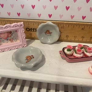 May include: A miniature set featuring a light pink ornate picture frame with a cherub image, two small scalloped ceramic bowls with cherub motifs, a pink rectangular tray with four rose-topped cupcakes, and one loose rose-topped cupcake.