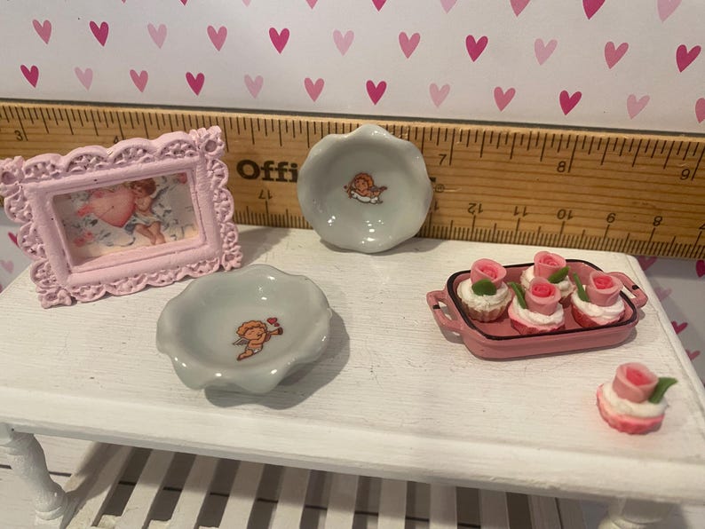May include: A miniature set featuring a light pink ornate picture frame with a cherub image, two small scalloped ceramic bowls with cherub motifs, a pink rectangular tray with four rose-topped cupcakes, and one loose rose-topped cupcake.