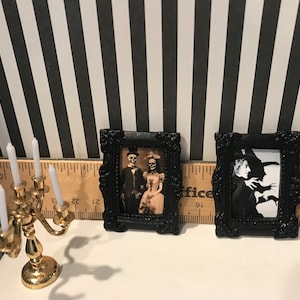May include: Miniature Halloween decor featuring two framed pictures with black ornate frames. One shows a skeleton couple, the other a witch. A gold candelabra with white candles sits nearby. The background is black and white striped.