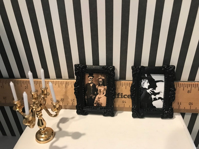 May include: Miniature Halloween decor featuring two framed pictures with black ornate frames. One shows a skeleton couple, the other a witch. A gold candelabra with white candles sits nearby. The background is black and white striped.