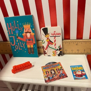 May include: Miniature Christmas-themed items on a white table. Includes a teal "Holly Jolly" card with a nutcracker, a "Toyland" card, a red "Season's Greetings" block, a record, and a "Woolworth's Toyland" card.