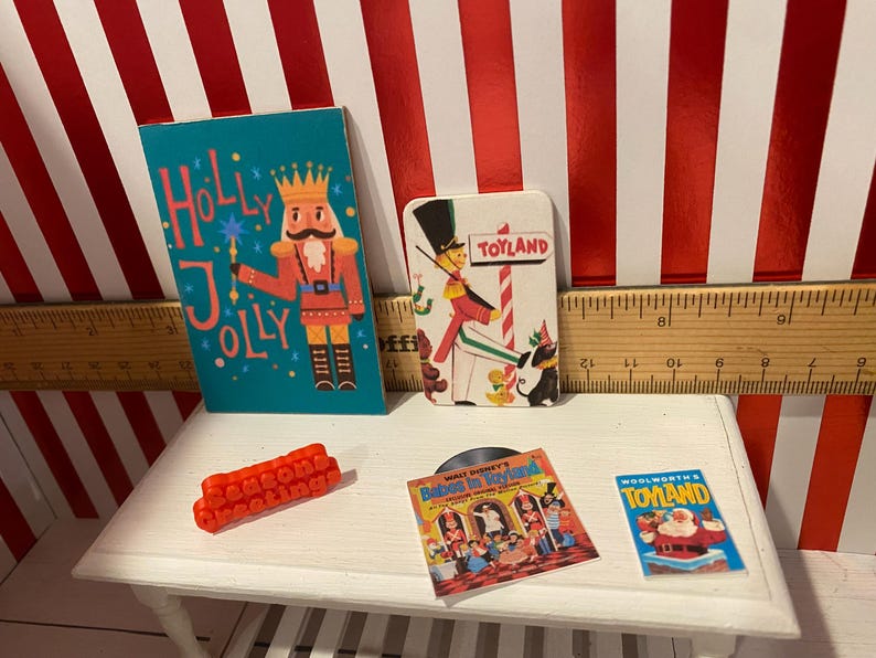 May include: Miniature Christmas-themed items on a white table. Includes a teal "Holly Jolly" card with a nutcracker, a "Toyland" card, a red "Season's Greetings" block, a record, and a "Woolworth's Toyland" card.