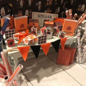Puede incluir: Una exposici&oacute;n en miniatura con tem&aacute;tica de Halloween con art&iacute;culos de papel naranja y negro, incluyendo cajas, pajitas y papel de envolver. La exposici&oacute;n presenta un calendario con la palabra "OCTUBRE" y una pancarta con tri&aacute;ngulos negros y naranjas.