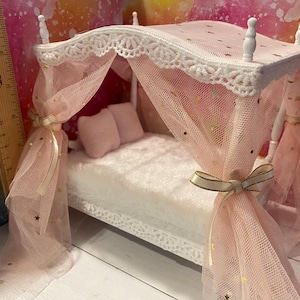 K&ouml;nnte beinhalten: Ein wei&szlig;es Puppenbett mit einem rosa Baldachin und goldenen Sternen. Der Baldachin ist aus T&uuml;ll und hat einen wei&szlig;en Spitzenbesatz. Das Bett hat eine wei&szlig;e Matratze und zwei rosa Kissen.