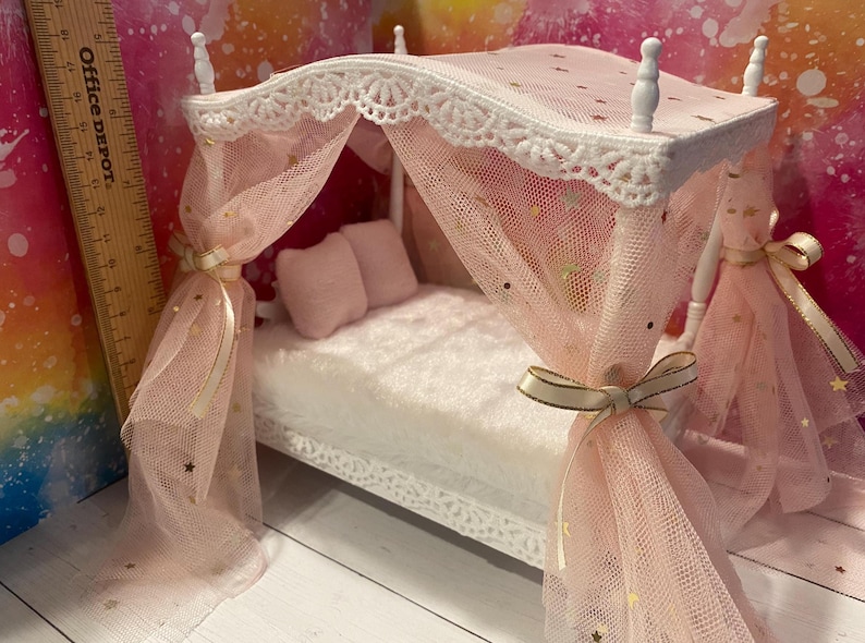K&ouml;nnte beinhalten: Ein wei&szlig;es Puppenbett mit einem rosa Baldachin und goldenen Sternen. Der Baldachin ist aus T&uuml;ll und hat einen wei&szlig;en Spitzenbesatz. Das Bett hat eine wei&szlig;e Matratze und zwei rosa Kissen.