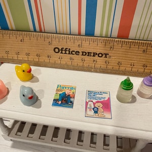 May include: Miniature baby-themed toys and accessories on a white table. Includes a yellow rubber duck, pink bird, blue bird, two baby bottles, and two magazines. A wooden ruler is in the background.