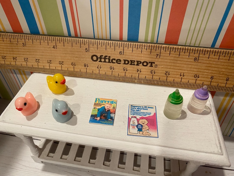 May include: Miniature baby-themed toys and accessories on a white table. Includes a yellow rubber duck, pink bird, blue bird, two baby bottles, and two magazines. A wooden ruler is in the background.