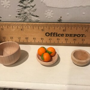 May include: Three miniature wooden bowls, one filled with three orange-colored miniature oranges, and two empty bowls. The bowls are on a white surface with a wooden ruler behind them. The ruler is labeled "Office Depot".