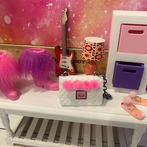 K&ouml;nnte beinhalten: Ein wei&szlig;es Puppenhaus-M&ouml;belset mit einer rosa und violetten Kommode, einer wei&szlig;en Handtasche mit rosa Bommeln, einer roten E-Gitarre und rosa und violetten flauschigen Stiefeln.