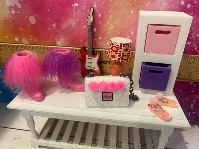 K&ouml;nnte beinhalten: Ein wei&szlig;es Puppenhaus-M&ouml;belset mit einer rosa und violetten Kommode, einer wei&szlig;en Handtasche mit rosa Bommeln, einer roten E-Gitarre und rosa und violetten flauschigen Stiefeln.