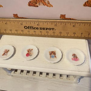 Pode incluir: Quatro pequenos pratos decorativos brancos com bordas onduladas, cada um com uma ilustra&ccedil;&atilde;o de tigre diferente. Os tigres s&atilde;o retratados com v&aacute;rios acess&oacute;rios, incluindo flores e uma m&aacute;scara azul. Os pratos s&atilde;o exibidos em uma prateleira branca.