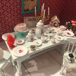 May include: Miniature Christmas-themed dining table setting with a cake, plates, cups, and a teapot. The table is set with a variety of festive items, including a snowman, a Santa Claus figure, and a gingerbread house. The scene is set against a red patterned background.