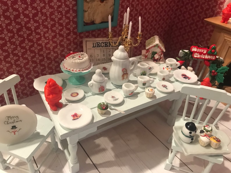 May include: Miniature Christmas-themed dining table setting with a cake, plates, cups, and a teapot. The table is set with a variety of festive items, including a snowman, a Santa Claus figure, and a gingerbread house. The scene is set against a red patterned background.