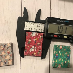 Puede incluir: Tres peque&ntilde;as cajas cuadradas con diferentes patrones navide&ntilde;os. Las cajas miden aproximadamente 2,57 cm de ancho. Las cajas est&aacute;n sobre una superficie de madera blanca.