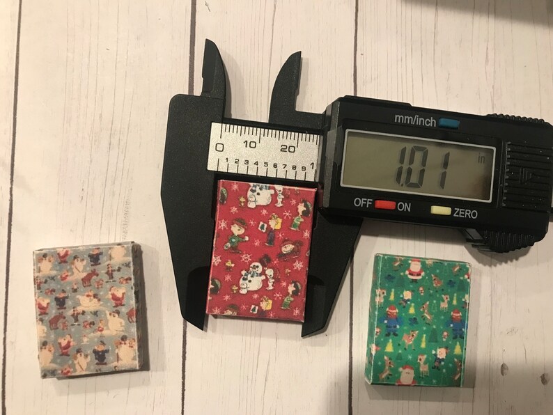 Puede incluir: Tres peque&ntilde;as cajas cuadradas con diferentes patrones navide&ntilde;os. Las cajas miden aproximadamente 2,57 cm de ancho. Las cajas est&aacute;n sobre una superficie de madera blanca.