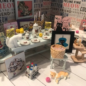 Könnte beinhalten: Eine Miniatur-Puppenhausszene mit einem weißen Tisch, der mit Teetassen und Untertassen, einer weißen Katzenfigur und einem Korb mit Kätzchen gedeckt ist. Es gibt mehrere gerahmte Bilder mit Katzenmotiven und einen Kratzbaum mit einer dreifarbigen Katzenfigur. Die Szene spielt auf einem weißen Holzboden.