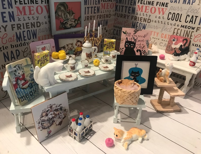 May include: A miniature dollhouse scene with a white table set with teacups and saucers, a white cat figurine, and a basket of kittens. There are several framed pictures with cat-themed designs and a cat scratching post with a calico cat figurine. The scene is set on a white wooden floor.