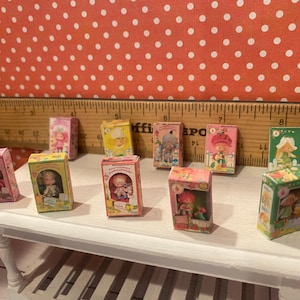 Op de afbeelding: Een verzameling miniatuur poppenboxen, elk met een andere karakterillustratie. De dozen zijn gerangschikt op een kleine witte tafel tegen een rode achtergrond met witte stippen. De dozen zijn in verschillende kleuren, waaronder roze, geel en groen.
