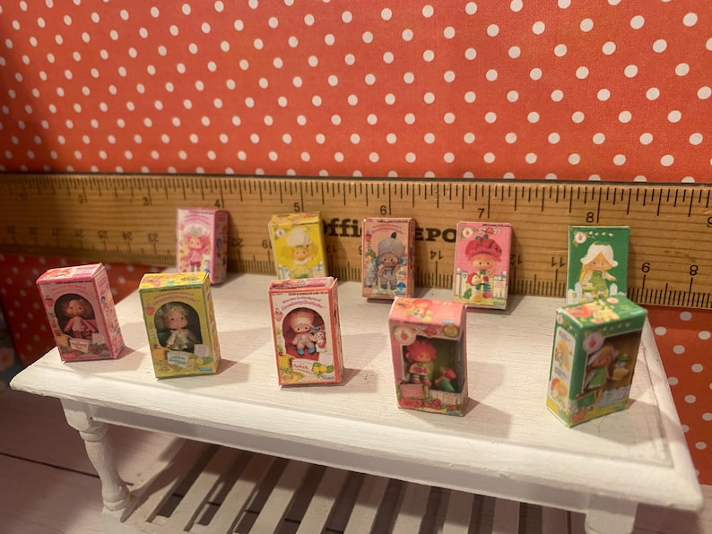 Op de afbeelding: Een verzameling miniatuur poppenboxen, elk met een andere karakterillustratie. De dozen zijn gerangschikt op een kleine witte tafel tegen een rode achtergrond met witte stippen. De dozen zijn in verschillende kleuren, waaronder roze, geel en groen.