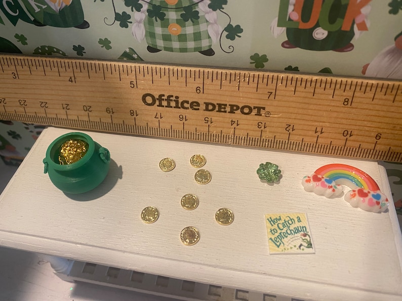 Puede incluir: Una olla verde llena de monedas de oro, un arco&iacute;ris, un tr&eacute;bol de cuatro hojas y un libro titulado "How to Catch a Leprechaun" sobre una superficie blanca. Tambi&eacute;n hay varias monedas de oro esparcidas alrededor de la olla.