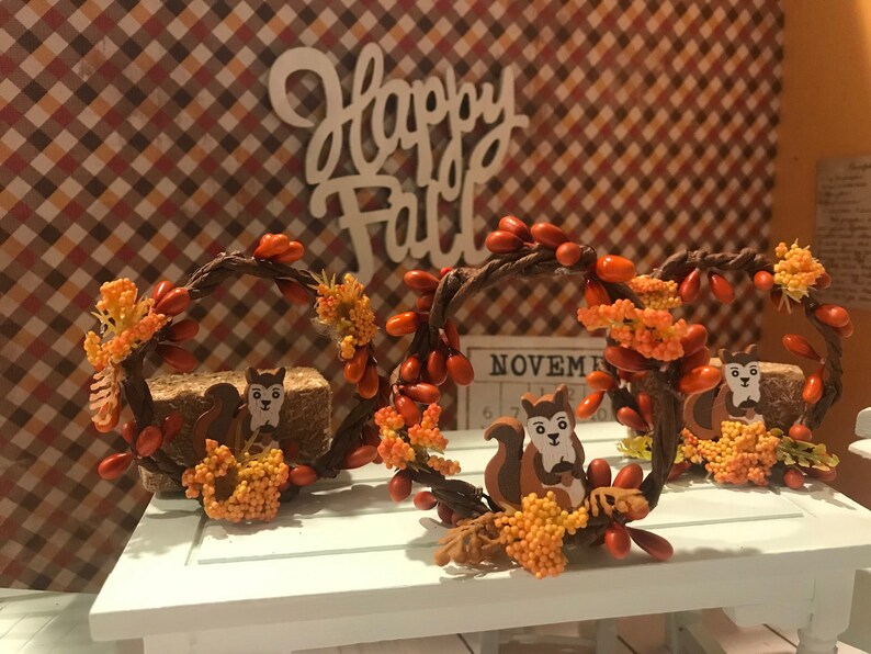 May include: Three fall-themed wreaths with brown twig bases, orange berries, and small orange and yellow flowers. Each wreath features a brown squirrel figurine with a white face.
