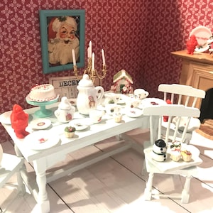 May include: A miniature Christmas dining scene with a white table set with plates, a cake, and a teapot. A framed Santa Claus portrait hangs on the red patterned wall. A plate on a chair reads "Merry Christmas".