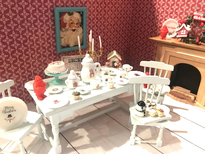 May include: A miniature Christmas dining scene with a white table set with plates, a cake, and a teapot. A framed Santa Claus portrait hangs on the red patterned wall. A plate on a chair reads "Merry Christmas".